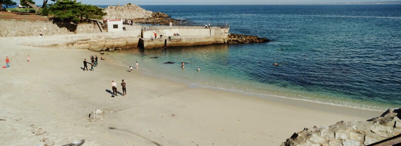 A sandy beach with people walking and playing near the water, bordered by stone walls and a small rocky pier, with clear blue ocean stretching to the horizon under a sunny sky.