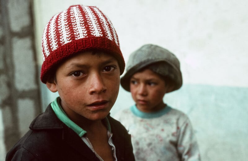 Two young boys stand near a wall. The boy in the foreground wears a red and white knit hat and looks at the camera, while the boy in the background wears a grey hat and looks away.