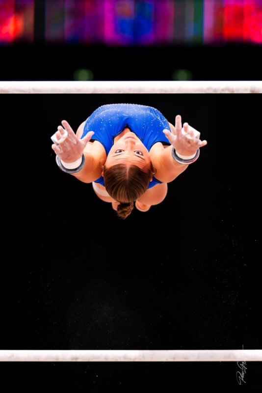 A gymnast in a blue leotard is seen mid-air, reaching upward between two uneven bars, with chalk on her hands and a focused expression, against a dark background with colorful lights above.