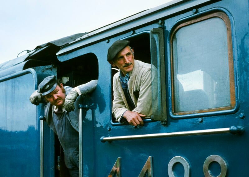 Two men wearing caps and work clothes lean out of the window and door of a blue steam locomotive, looking ahead. The side of the train displays the number 4498.