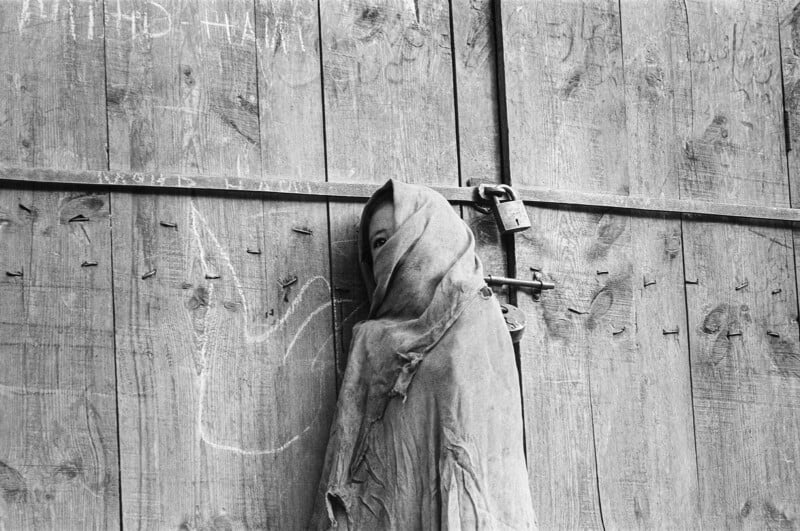 A person wearing a tattered, hooded cloth stands against a weathered wooden door secured with a padlock, with only one eye visible. Faint chalk drawings and writings appear on the door.