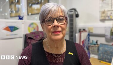 A close-up image of Cherry Cantrell sitting at the desk in her office. She is an older woman, with a purple hue in her short hair. She is wearing  apair of circular glasses, a silver necklace, as well as a purple lace top and black waistcoat. She has a yellow 'Aware NI' pin on her lapel. She is looking at the camera and smiling. Behind her are posters and lights on the wall of her office, but they are blurred.