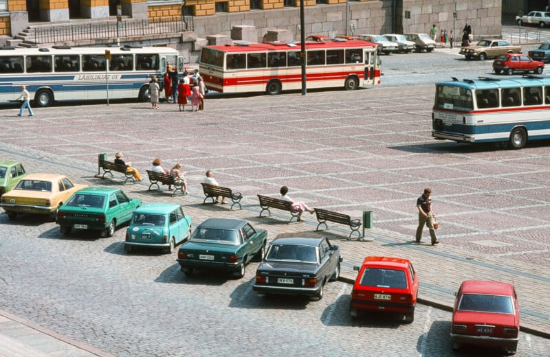 An open square with parked cars and several benches, some occupied by people. Three buses are parked in the background, and a few individuals are walking or standing around the square. The ground is paved with patterned stones.