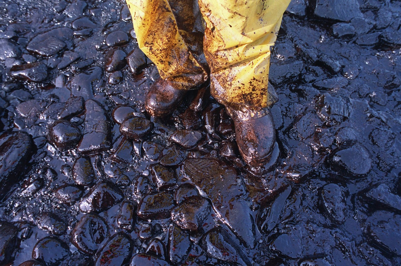 Thick crude oil, dark brownish black, sticks to the rocks and covers the boots and yellow pants of a local fisherman