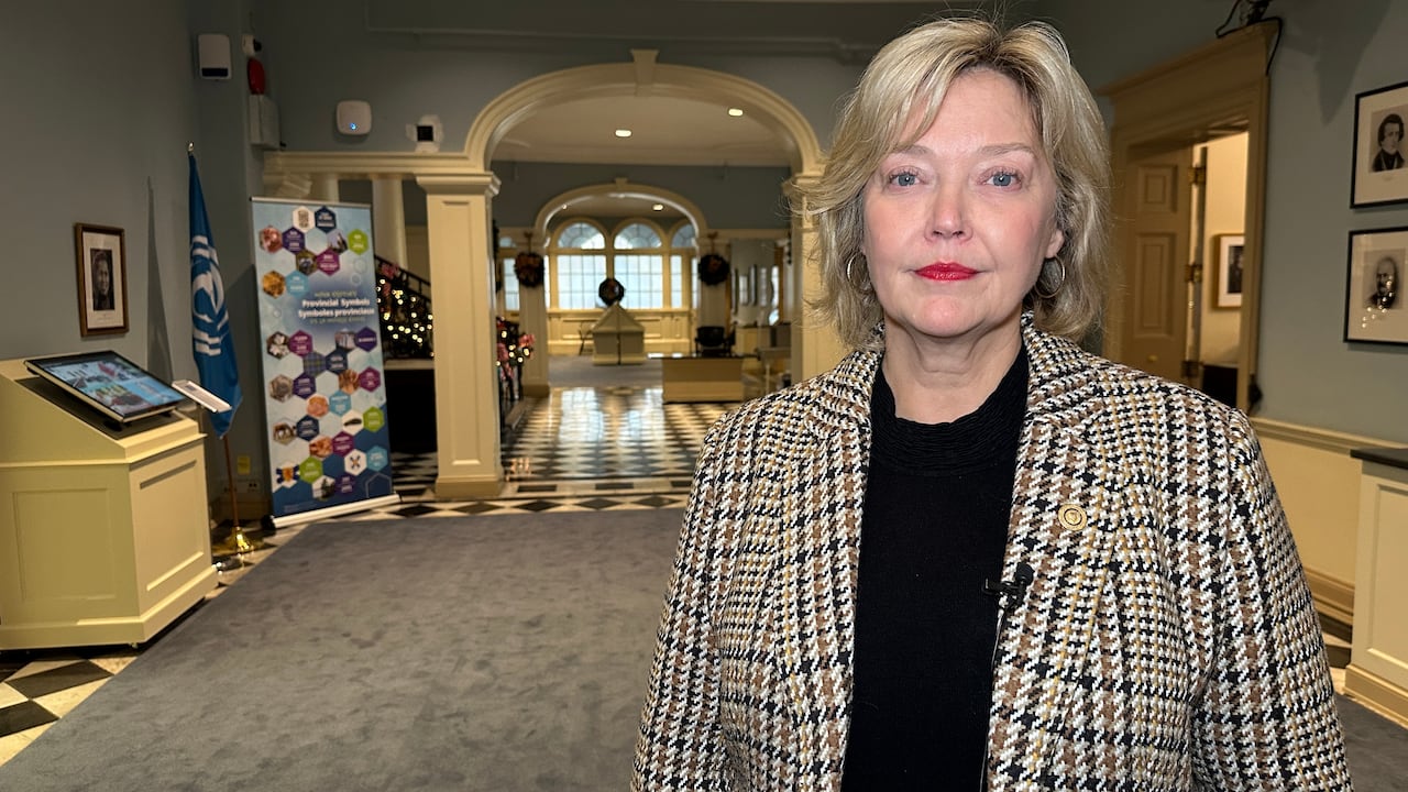A woman in a checked jacket and black sweater is looking at the camera with an archway in the background.