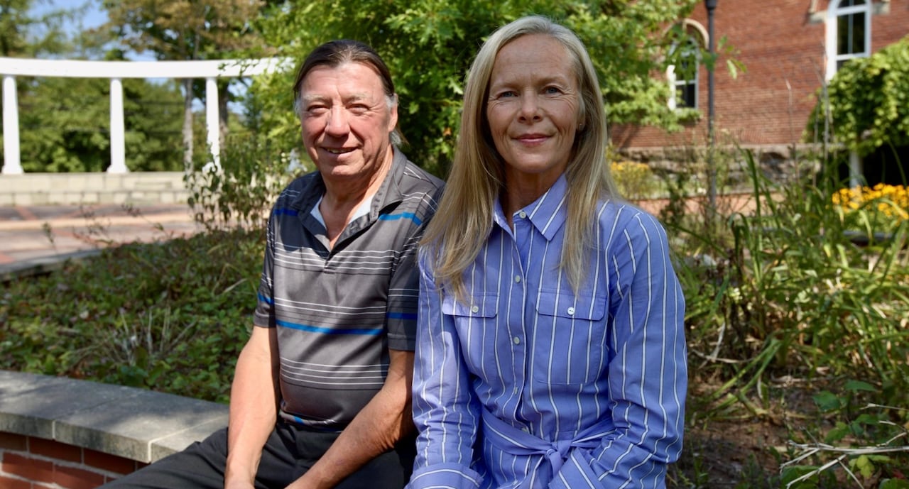 Two people sit together on a bench outside, in front of a brick building and some shrubbery.