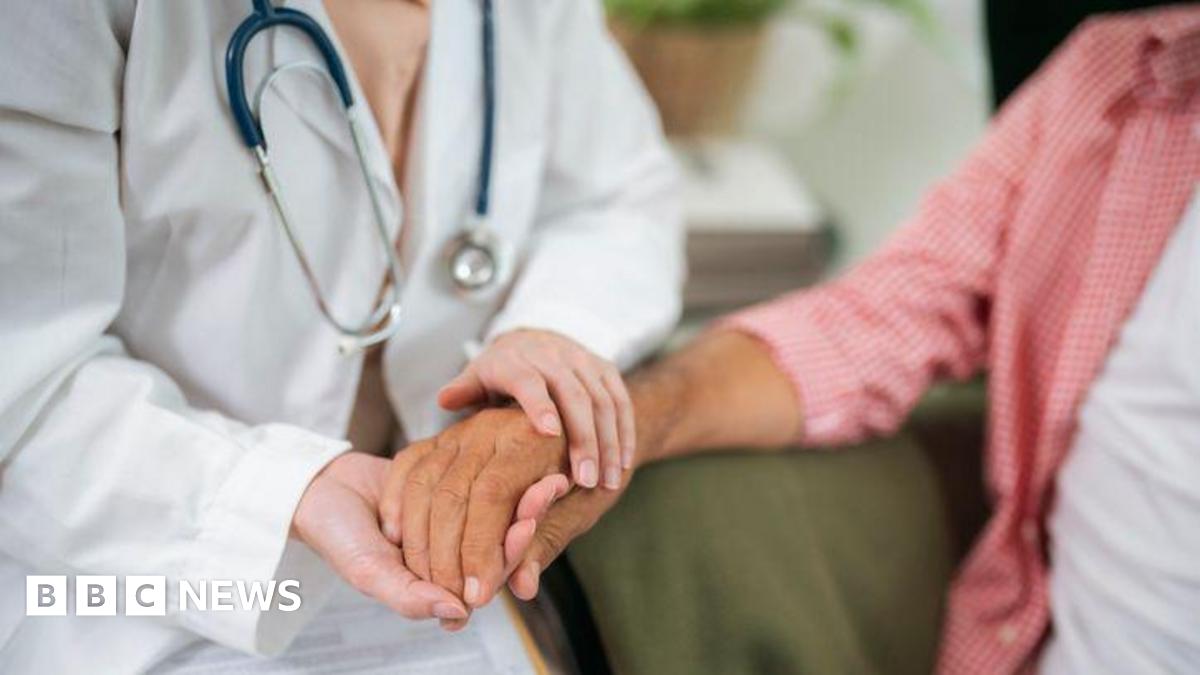 A doctor holds a woman's hand in a consultation. The doctor has a white coat and a stethoscope.