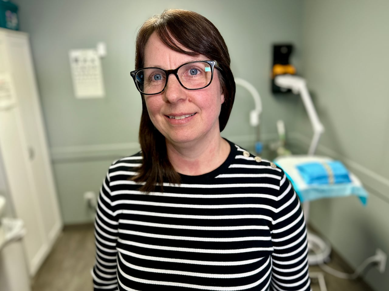 A woman with glasses smiles at the camera in a medical clinic room.