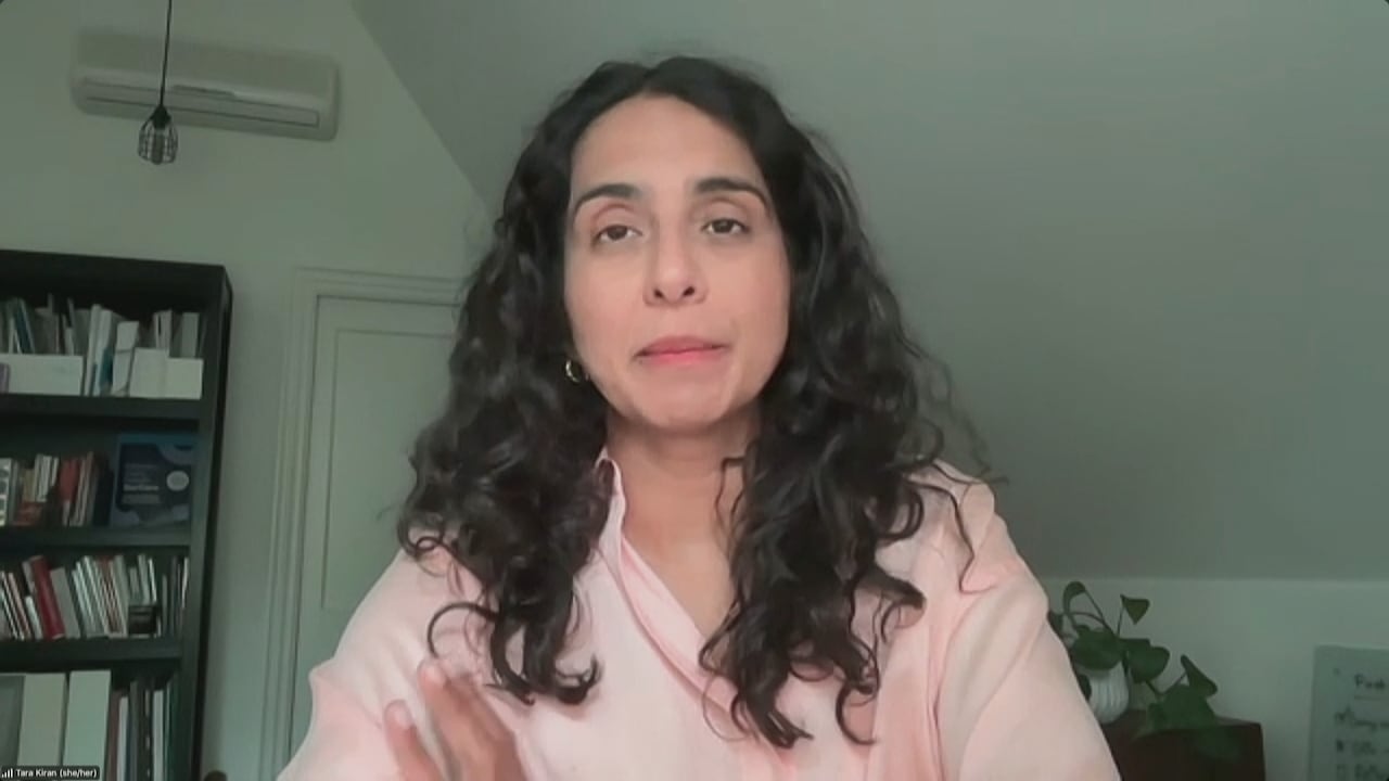 Woman with long curly black hair wearing a pink blouse as she sits in front of a bookcase. 