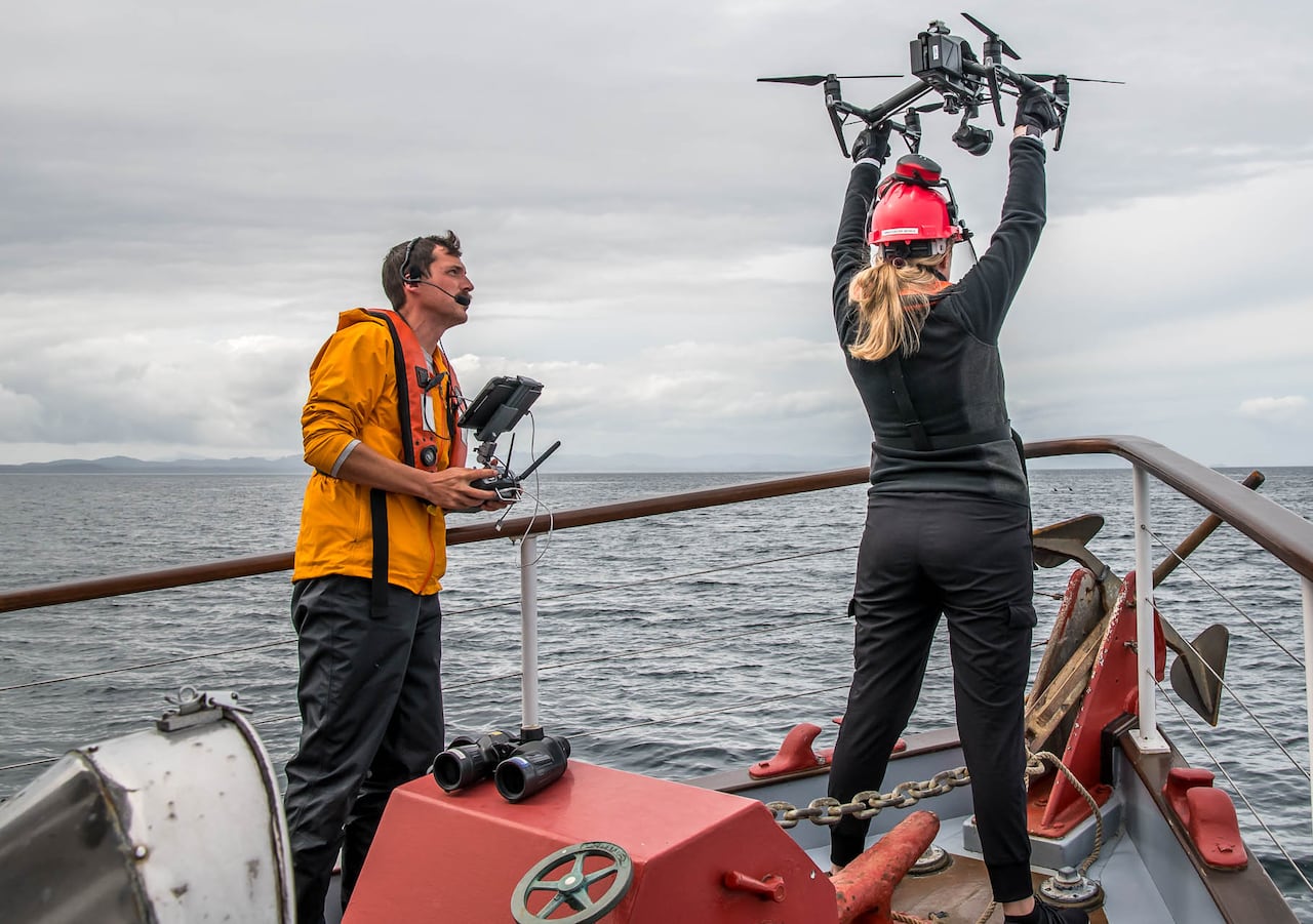Two people operate a drone at the tip of a boat.
