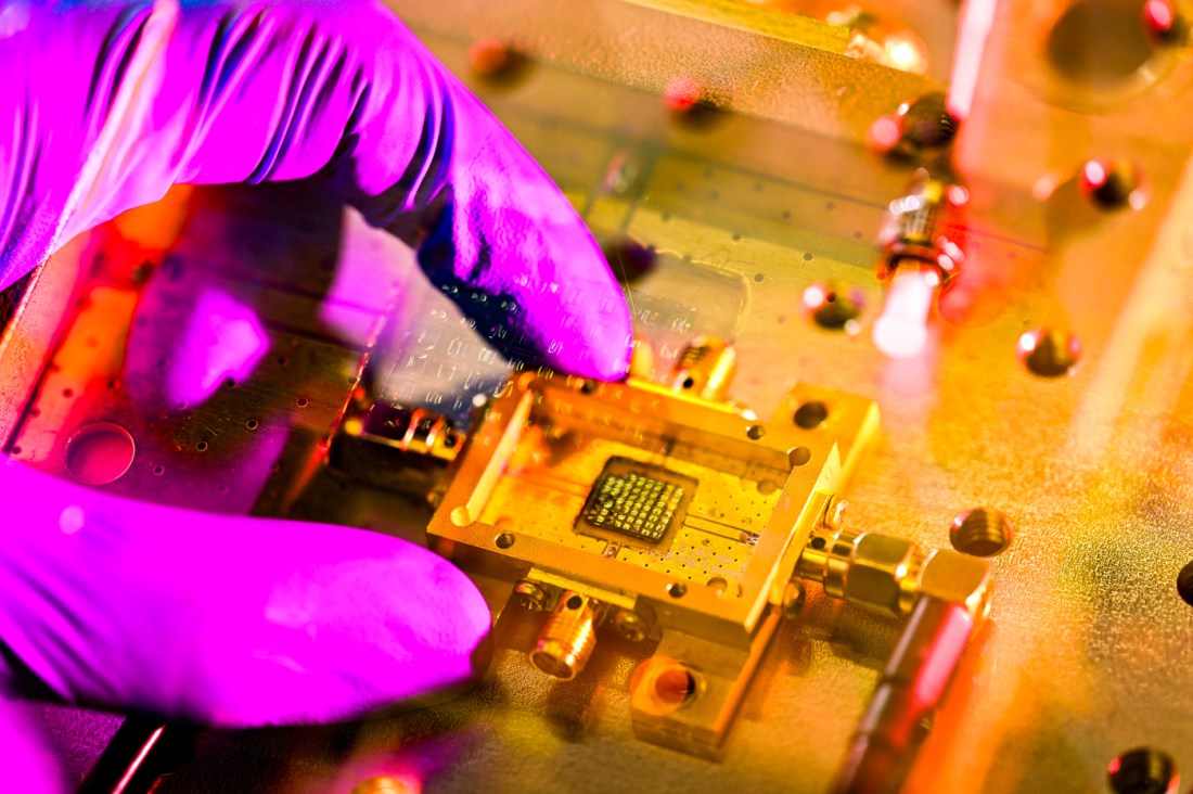 A hand with a blue latex glove, turned purple by the camera light, manipulates a sensor in a gold housing with a computer chip at its center.