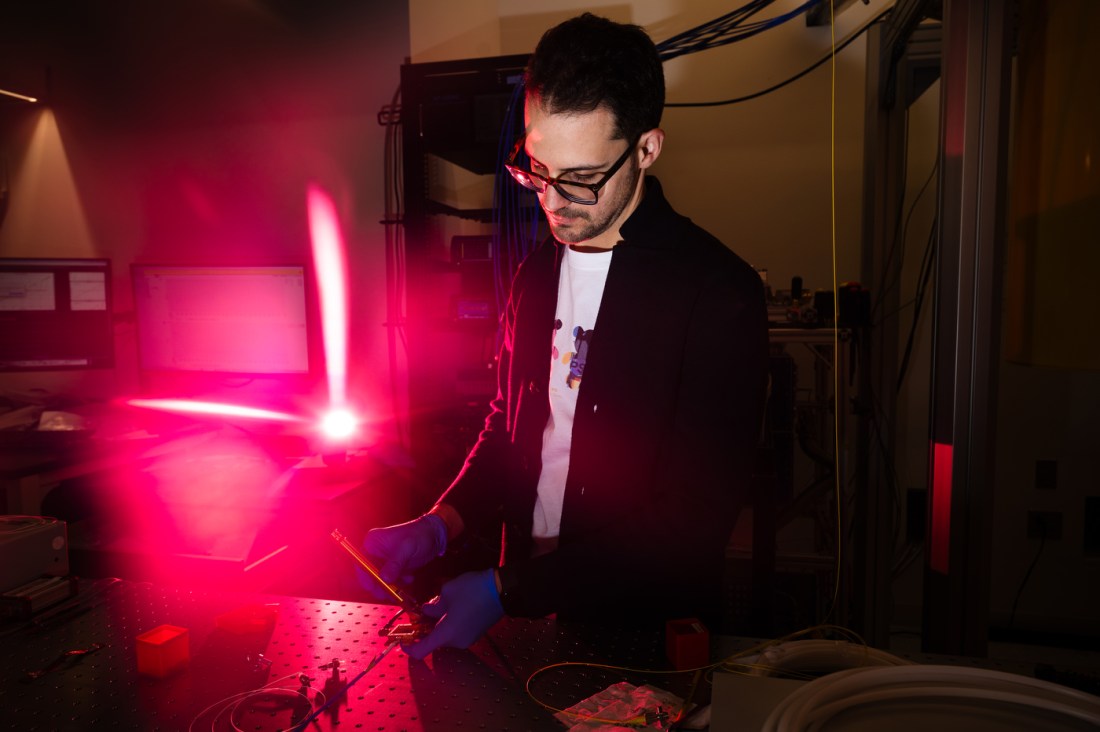 A man wearing glasses stands next to a table manipulating a sensor. A red light illuminates him from behind.