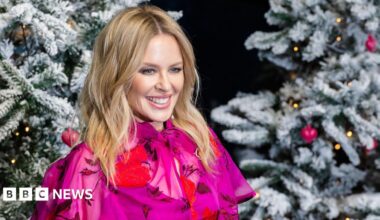 Kylie Minogue poses in a pink shirt next to a pair of Christmas Trees