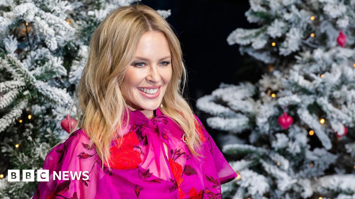 Kylie Minogue poses in a pink shirt next to a pair of Christmas Trees
