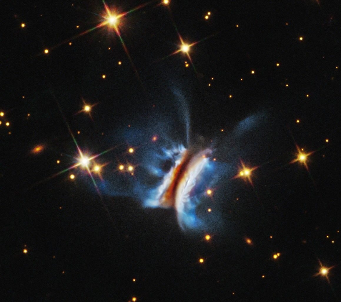 Near the center is an object that resembles an edge-on view of a hamburger. There is a diagonal dark strip (the meat patty) of dust, running from 1 o'clock to 7 o'clock, that obscures a central star. Curving away from either side of the dark strip are glowing white clouds (the buns) where dust is reflecting starlight. Bright blue finger-like wisps of material extend far above and below the dark center plane. A few dozen stars, some with four diffraction spikes, are scattered on the black background of space.