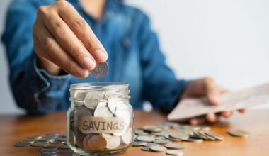 Woman hand is putting a coin into a glass bottle next to a pile of coins on a brown wooden table