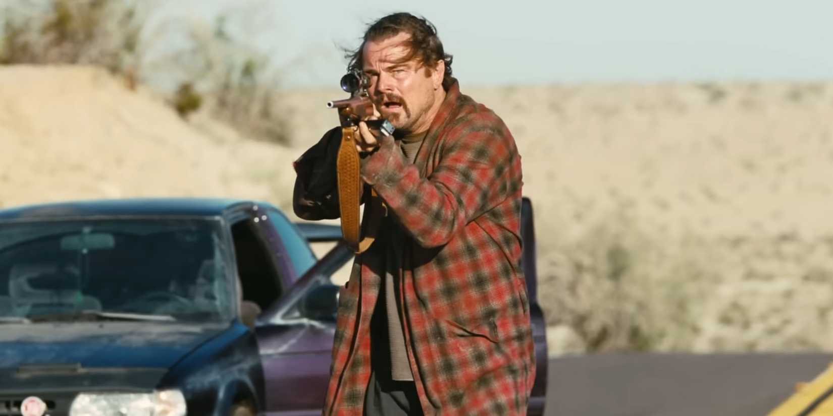 Leonardo DiCaprio aims a weapon while walking near his stopped car in One Battle After Another