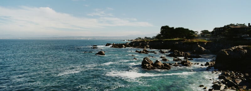 Rocky coastline with waves crashing against the rocks under a clear sky, distant mountains visible on the horizon, and houses atop the cliff on the right side of the image.