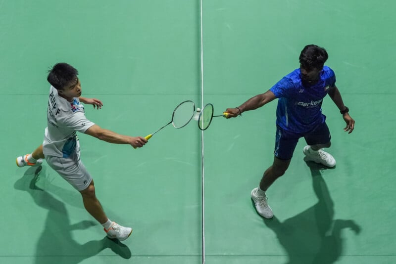 Two badminton players on a green court touch their racquets at the net, one in a white outfit and the other in blue, as seen from an overhead perspective.