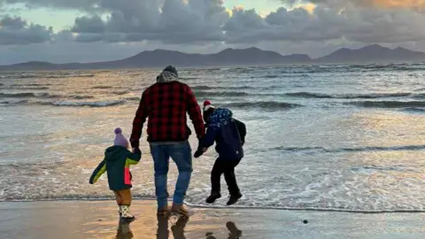 Aled Edwards A beach at sunset. A man stood on the sand is facing away from the camera, with each hand holding that of a child wrapped up warm - one boy and one girl.