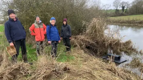 RTCT Four volunteers part of the team that deploys, monitors and records data from rafts along the River Thame. They are posing for an image by the river on an overcast day.
