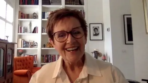 A woman with short hair and glasses smiles into the camera, with an orange chair and a floor-to-ceiling bookcase behind her