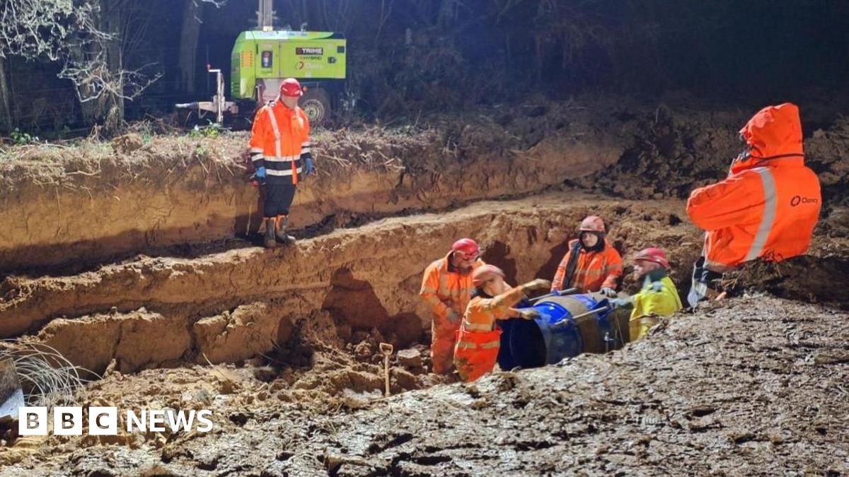Four men in high-vis orange clothing stand next to a large pipe in an excavated trench. It is dark but the work site is brightly lit. Two other men are stood higher up the banks of the trench.