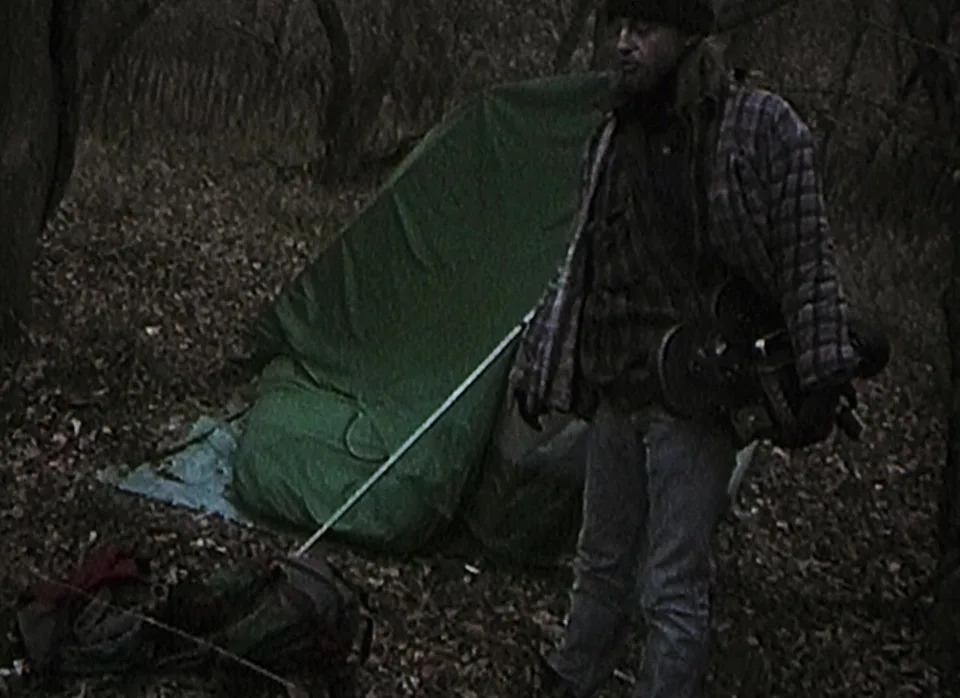 Person standing near a tent in a wooded area, holding a guitar, wearing a flannel shirt and cap. The ground is covered in fallen leaves
