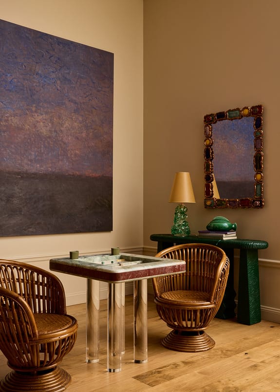 Seating area with wicker chairs, a glass table, abstract wall art, a decorative mirror, a lamp, and a textured console table.