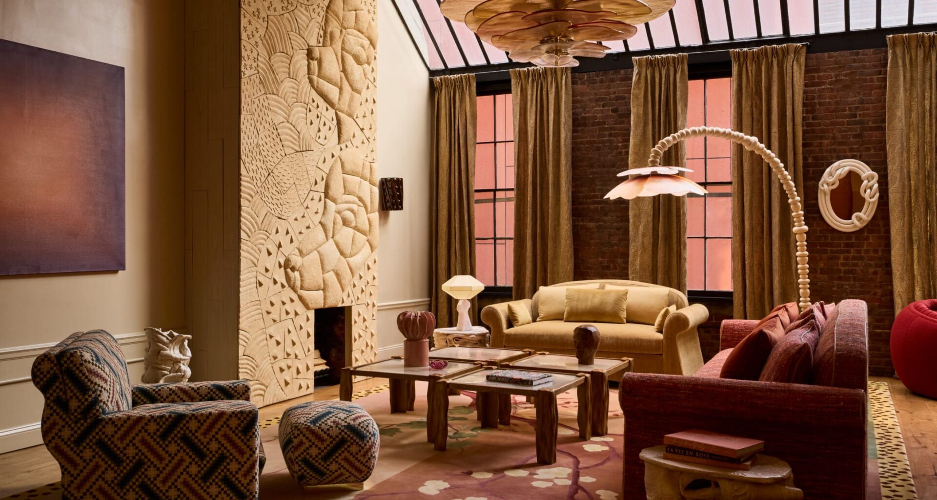 Eclectic living room with patterned furniture, carved wall art, large chandelier, and tall windows with brown curtains.