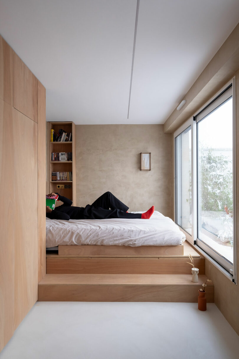 Person lies on a raised bed in a minimalist bedroom with wood accents, built-in shelving, large window, and neutral-toned walls.