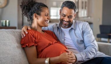 Pregnant woman on couch with man, both smiling and touching her belly.
