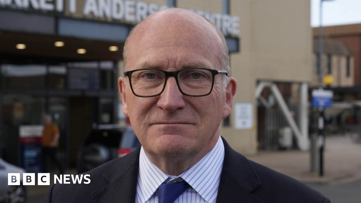Nick Hulme looks at the camera with a serious expression outside Colchester Hospital. He wears a black suit with a white shirt underneath that has blue stripes on it. He also wears a navy tie.