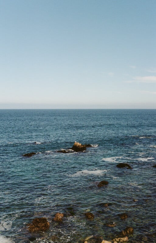 Clear blue ocean with gentle waves and scattered rocks in the water under a bright, cloudless sky. The distant horizon meets the calm sea, creating a peaceful and open coastal scene.