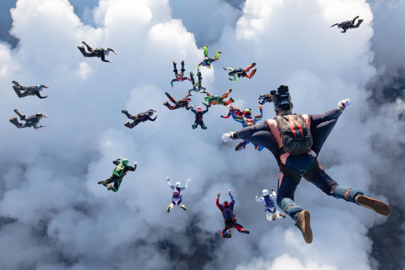 A group of skydivers in colorful suits are free-falling through the sky above the clouds, some holding hands in formation, while one skydiver with a camera is in the foreground facing the group.
