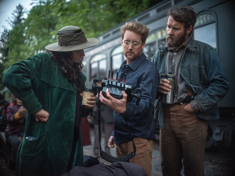 Three people stand outdoors near a vintage train; one holds a device with antennas while the others observe. They wear retro-style clothing and appear to be discussing something technical. Trees and train cars are visible in the background.