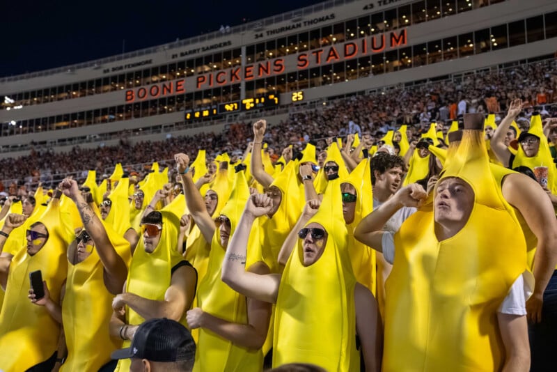 A large group of fans in banana costumes cheer in the stands at Boone Pickens Stadium during a nighttime sports event. The crowd is energetic and the stadium is packed.