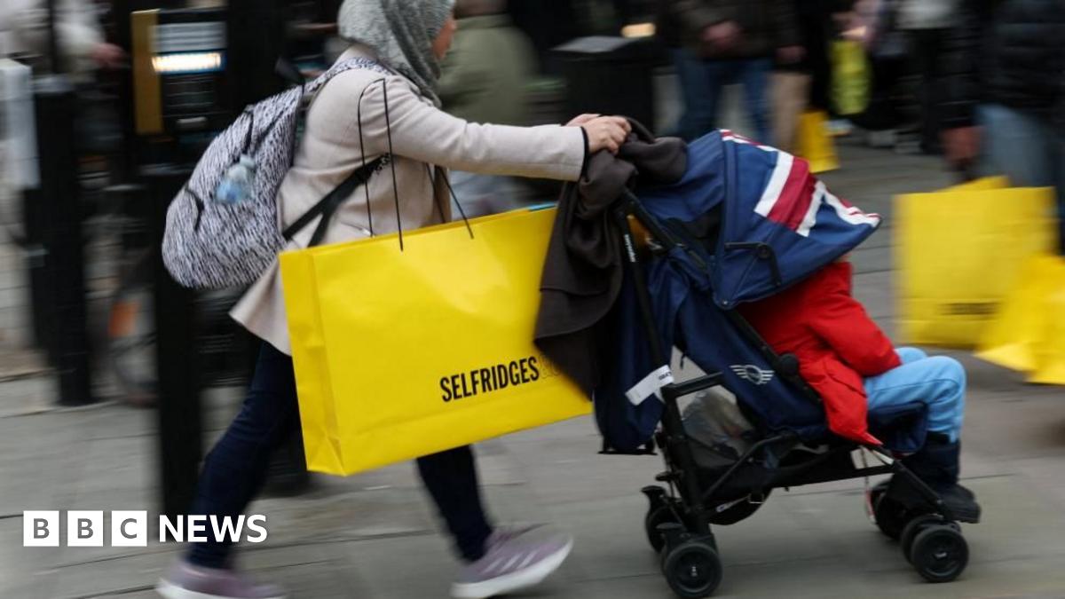 A woman wearing a hijab pushes a pram while carrying a large yellow Selfridges bag down the street.
