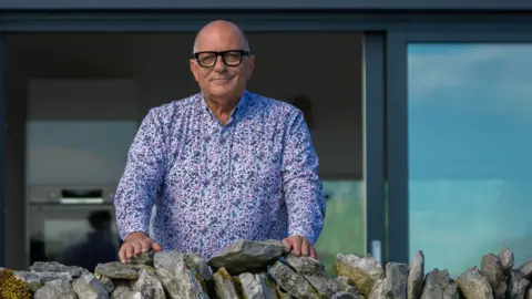 Animo TV 2025 Hugh Wallace standing behind a stone wall with his hands placed on the wall. He is bald, wearing glasses and a purple floral shirt. A glass door is behind him.