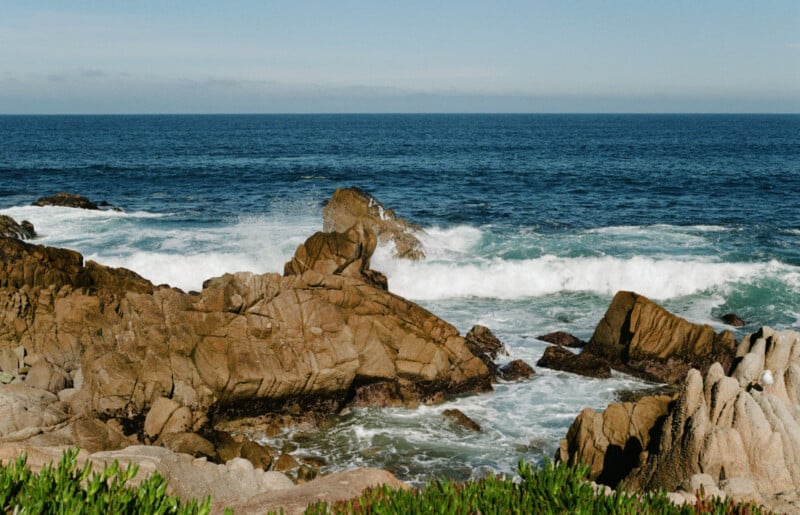 Waves crash against large, jagged rocks along a rugged coastline under a clear blue sky, with some green vegetation visible in the foreground.