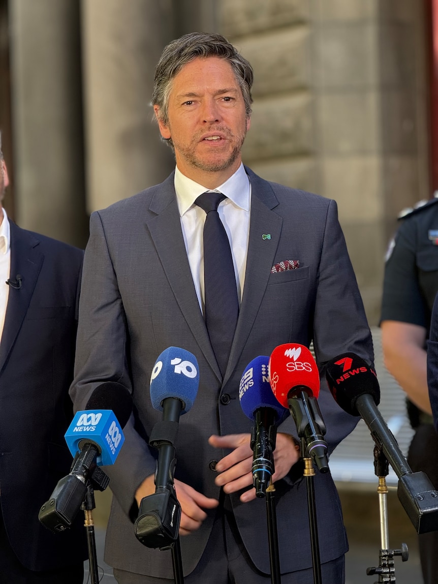 A man with grey hair in a grey suit, white shirt and black tie stands behind microphones in front of a sandstone building.
