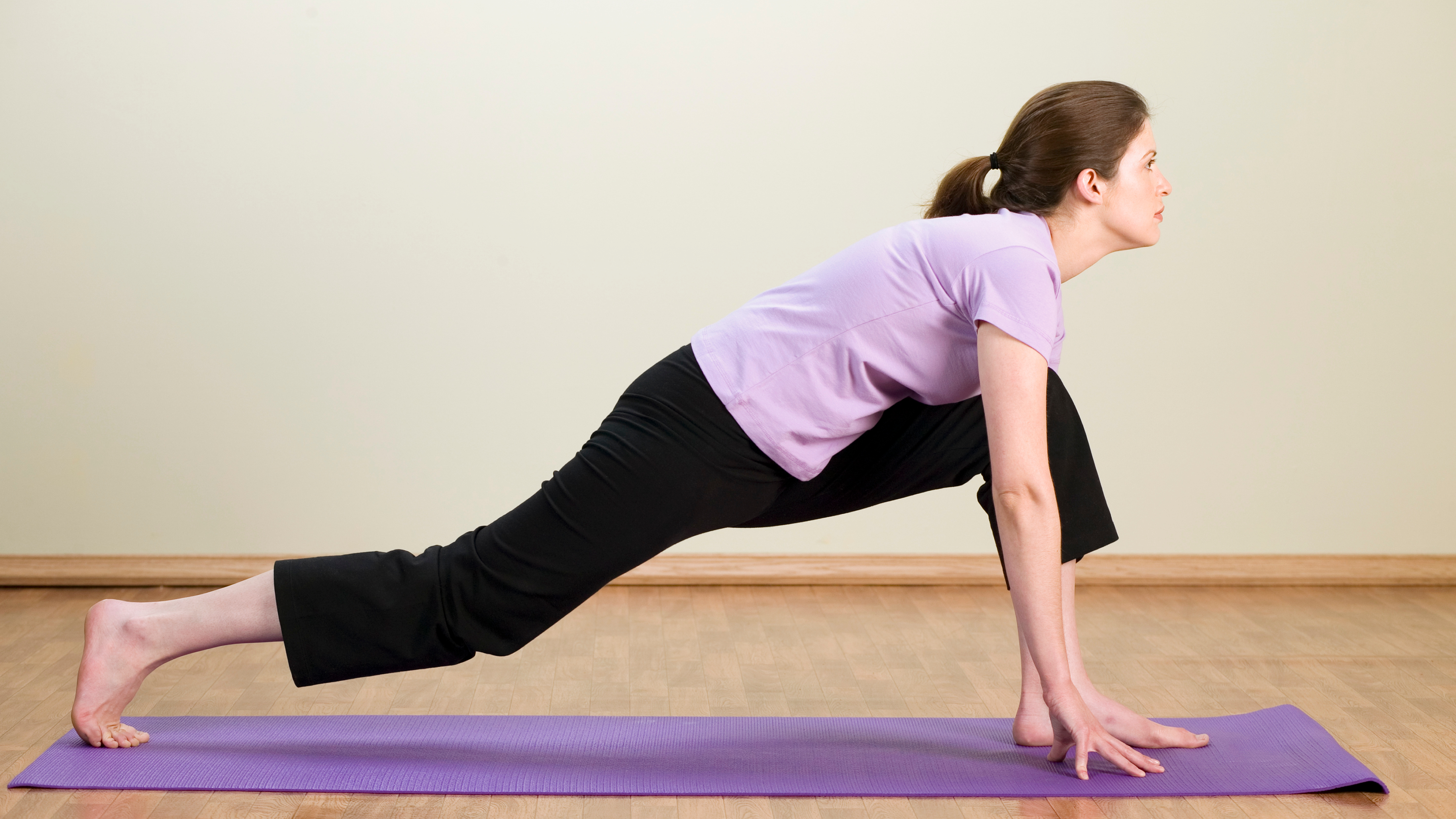 A woman performs a low lunge on an exercise mat. Her left foot is forward, left knee bent, and her right leg is straightened behind her. Her hands are on the floor either side of her front left foot.
