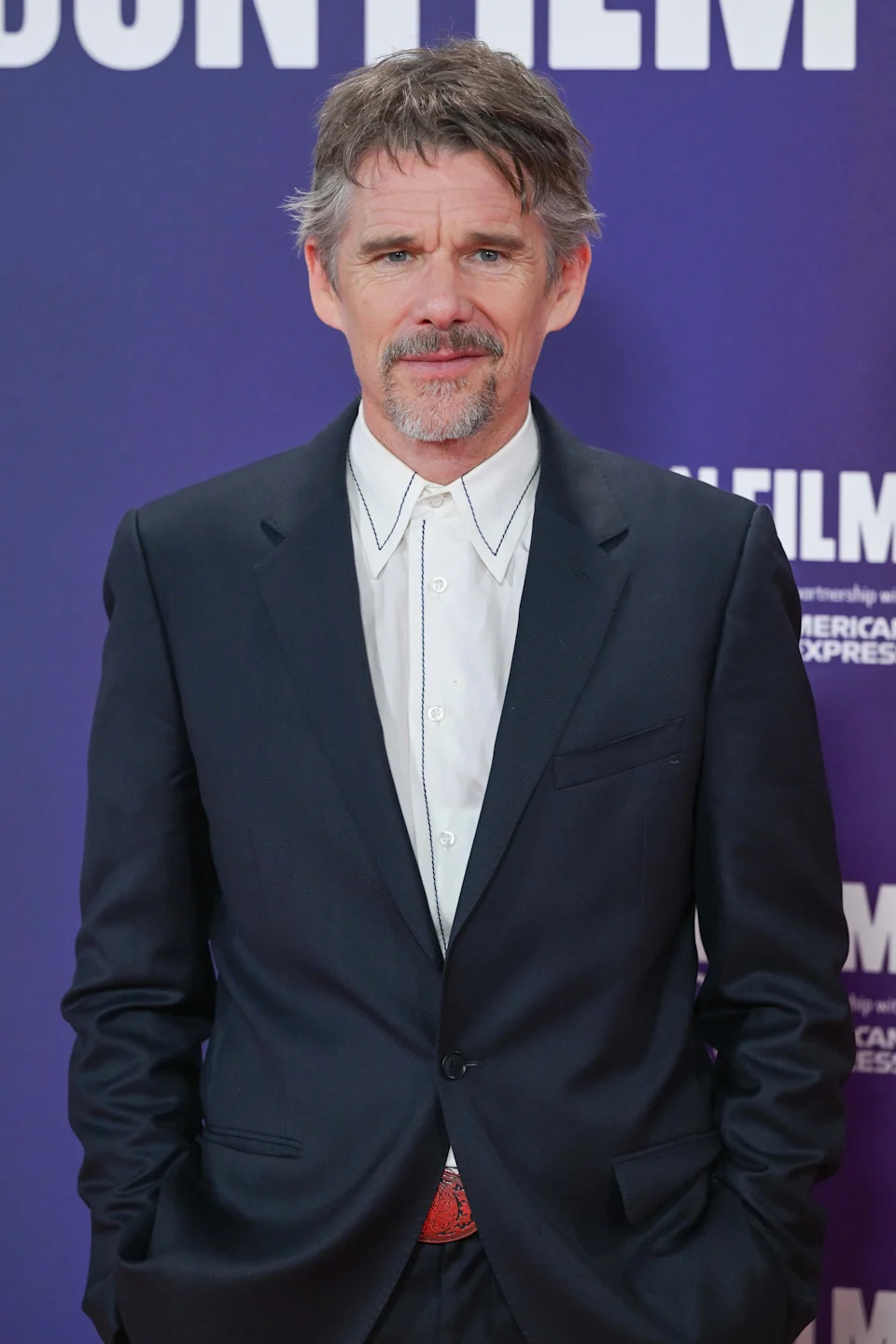 Person in a tailored suit with hands in pockets, standing against a backdrop with text, likely at a celebrity event