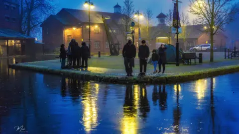 Mark Pemberton Seven people looking at a frozen canal. The canal reflects blue and yellow colours