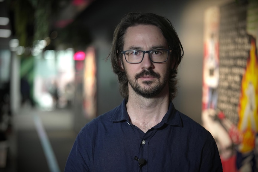 A man in a navy collared shirt with a beard and glasses stands in a colourful, modern office corridor.