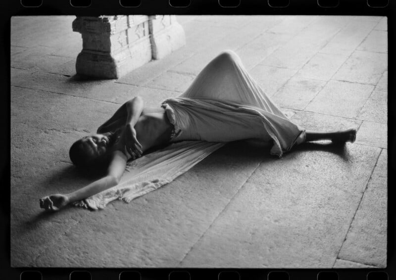 A person draped in light fabric lies on a stone floor with one arm extended and eyes closed, in a relaxed pose. The setting appears to be a spacious, sunlit area with stone columns nearby. The image is in black and white.