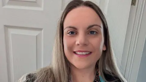 Katie Graden A selfie of a woman with blonde hair smiling. She is standing indoors in front of a white door.