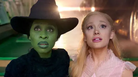 Universal Cynthia Erivo and Ariana Grande stare into the camera dressed as their Wicked characters Elphaba and Glinda, in a scene from the film Wicked. Elphaba wears a black outfit and black witch's hat and is painted green. While Glinda has blonde hair and wears a pink dress.