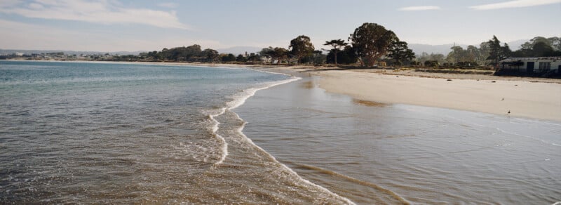 A calm beach scene with gentle waves washing onto the sandy shore, bordered by trees and distant low hills under a partly cloudy sky. A building is visible on the right side near the edge of the beach.
