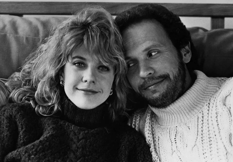 Two people seated closely, smiling warmly. The person on the left wears a turtleneck, and the person on the right wears a knit sweater